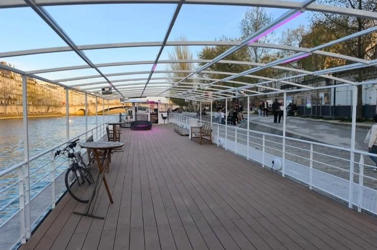 Le pont couvert d’une verrière, avec tables hautes et bancs, le long du quai.